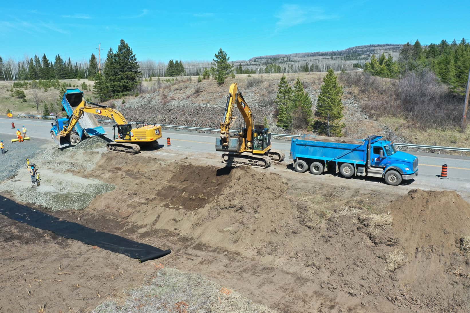 Two blue dump trucks and two yellow excavators at work moving sand at a highway construction site. LH North workers are compacting sand and laying pipes.