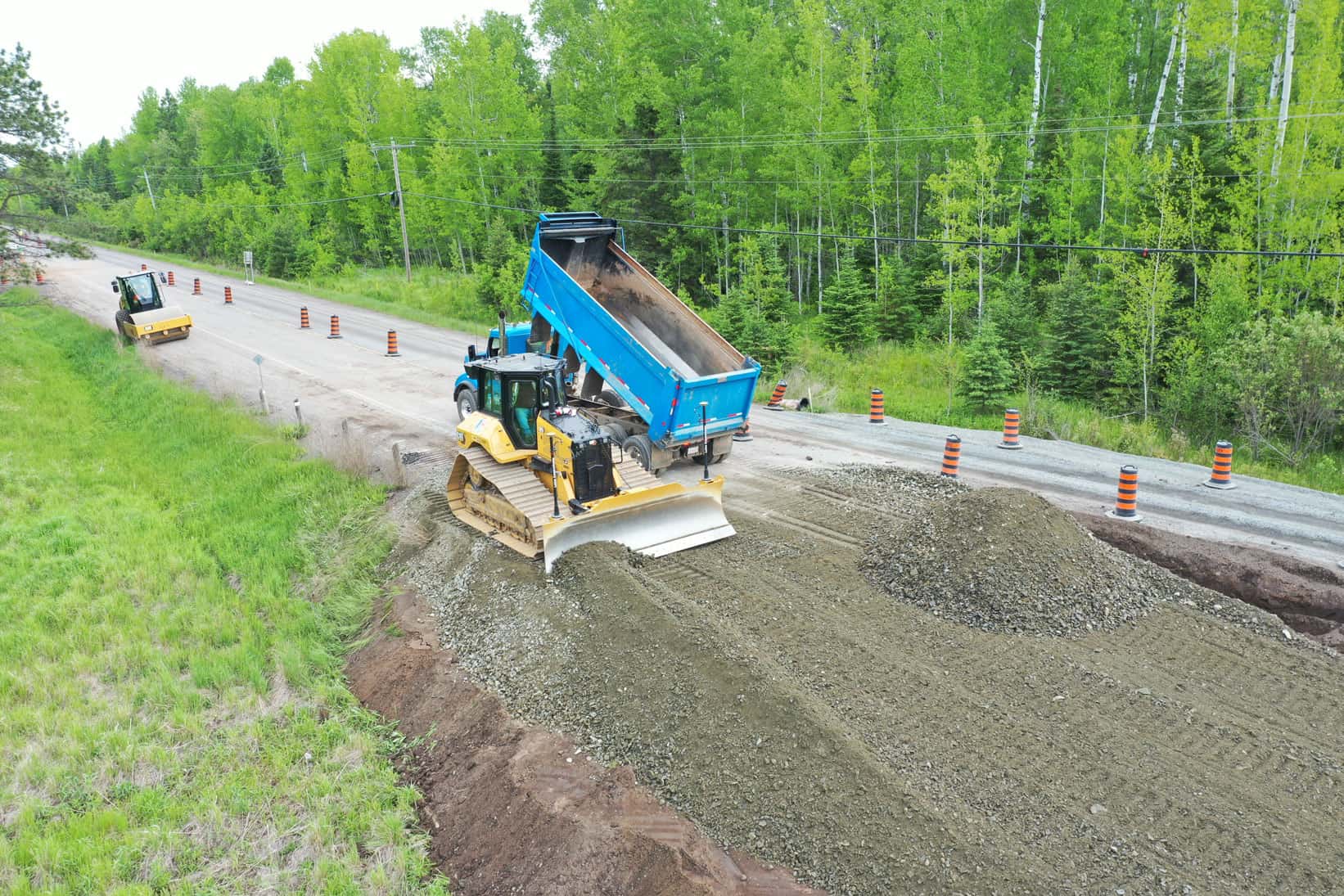 A bulldozer and a blue dump truck are hard at work on a LH North highway reconstruction project. In the forefront is gravel and sand, whereas the forest is impossibly green in the background.