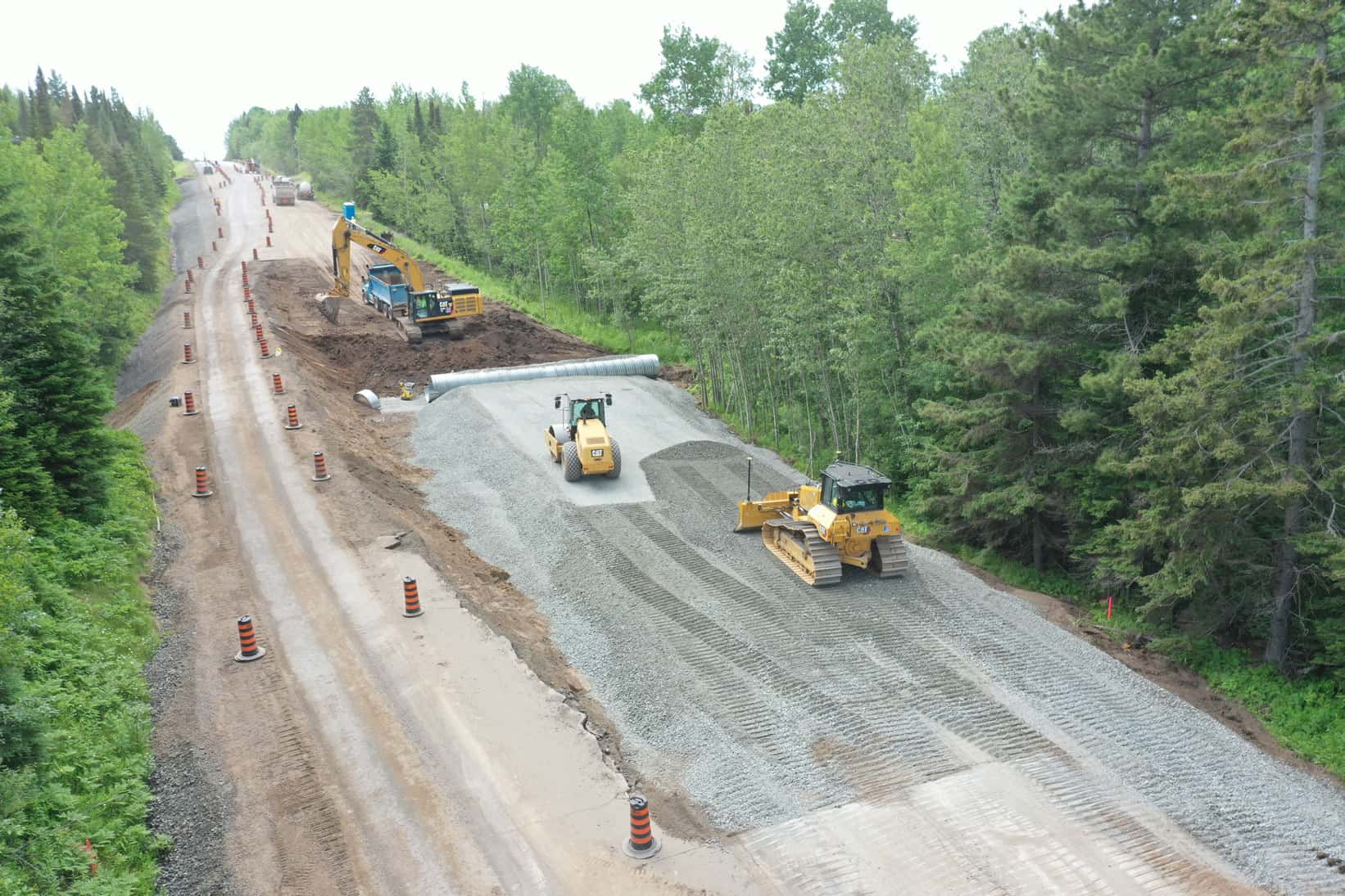A drone view of an LH North construction project of a highway. A bulldozer and compacter sit in the forefront of the image, whereas an excavator moves sand at the back.