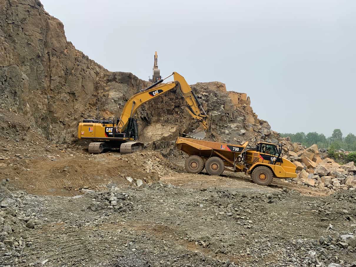 An CAT excavator and dump truck sit against a rocky backdrop in this image. They are moving rocks and rubble in a highway reconstruction project by LH North.