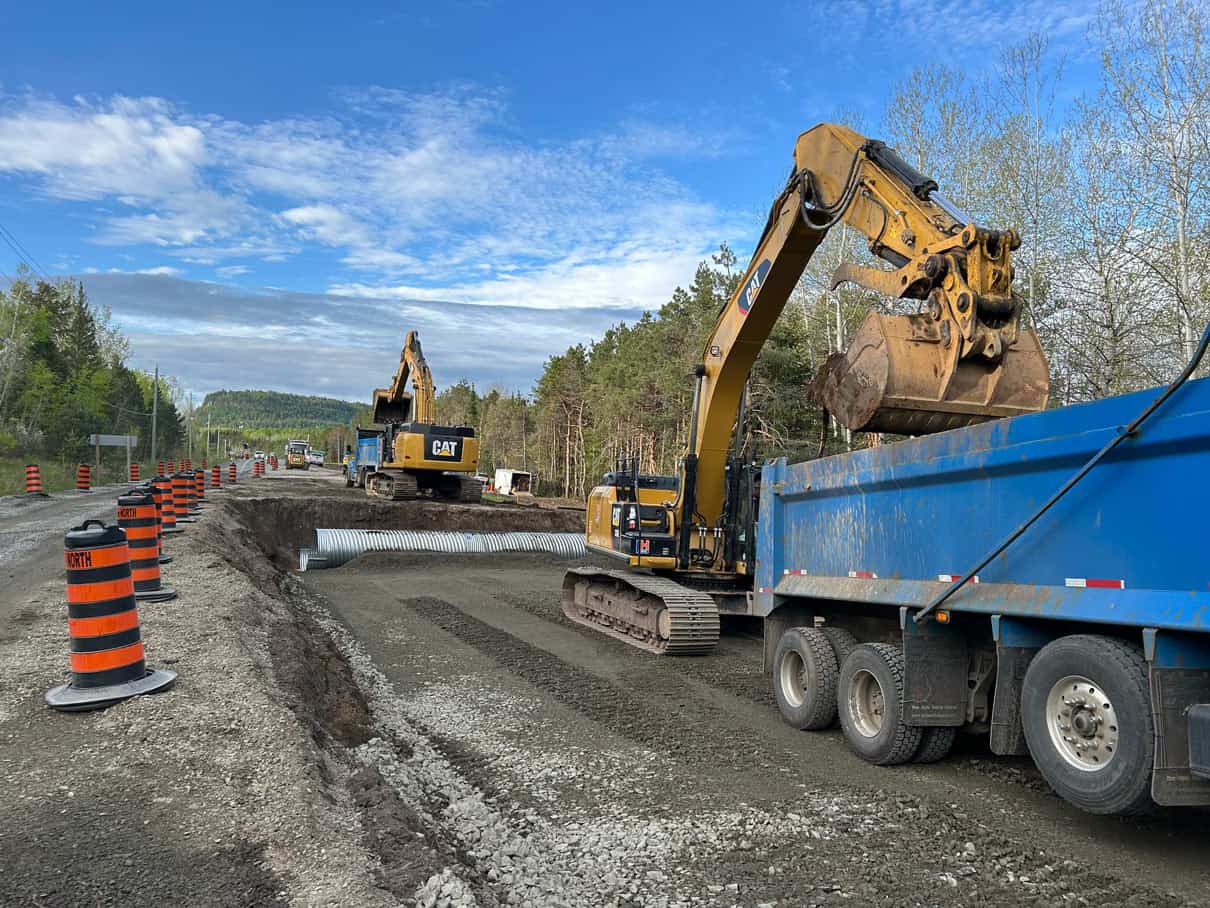 A highway construction site. Two LH excavators work to fill dump trucks while black and red safety cones tell motorists where to go.
