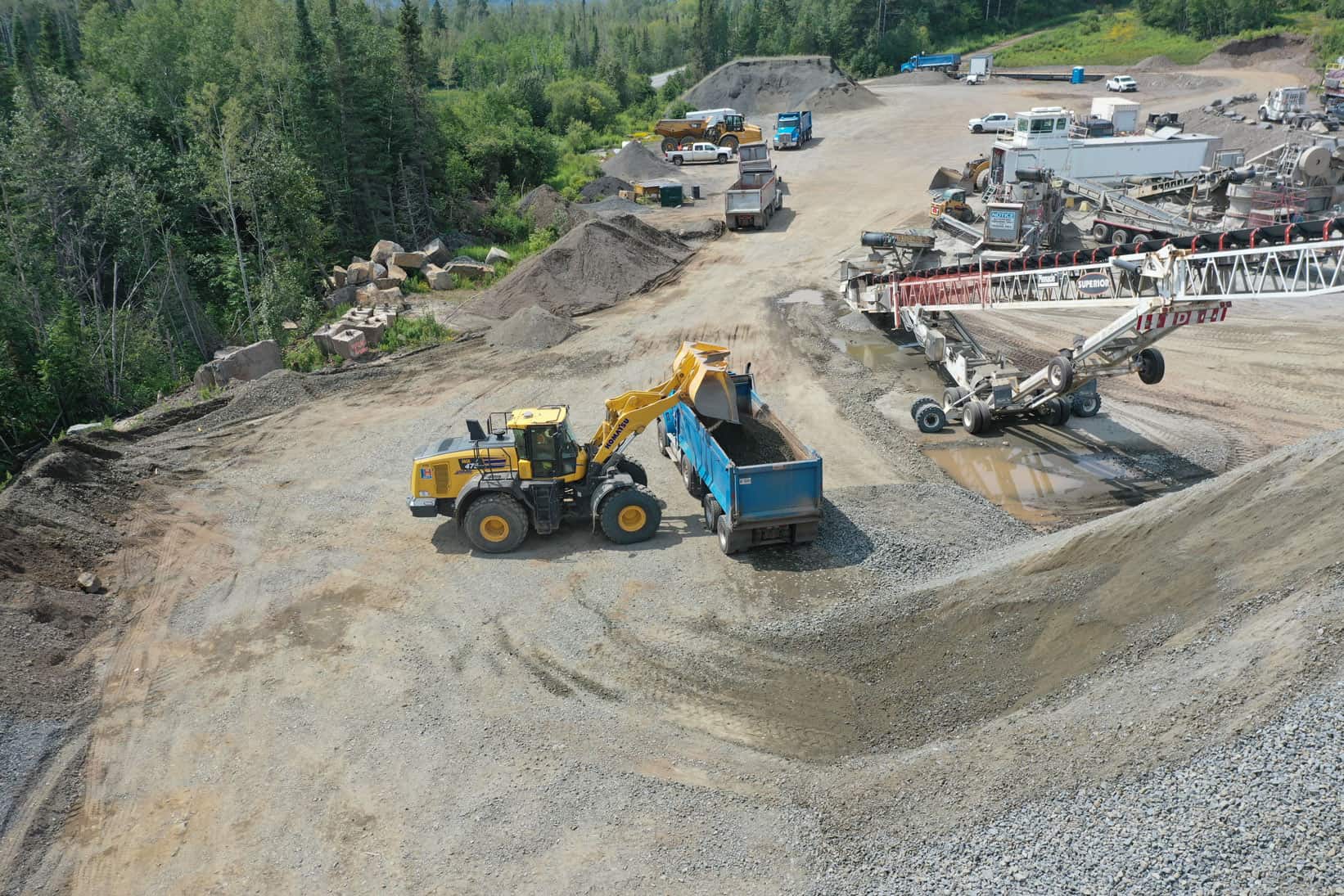 An LH excavator dumps gravel in a blue dump truck. On the right is an industrial crane, ready for when it is needed.